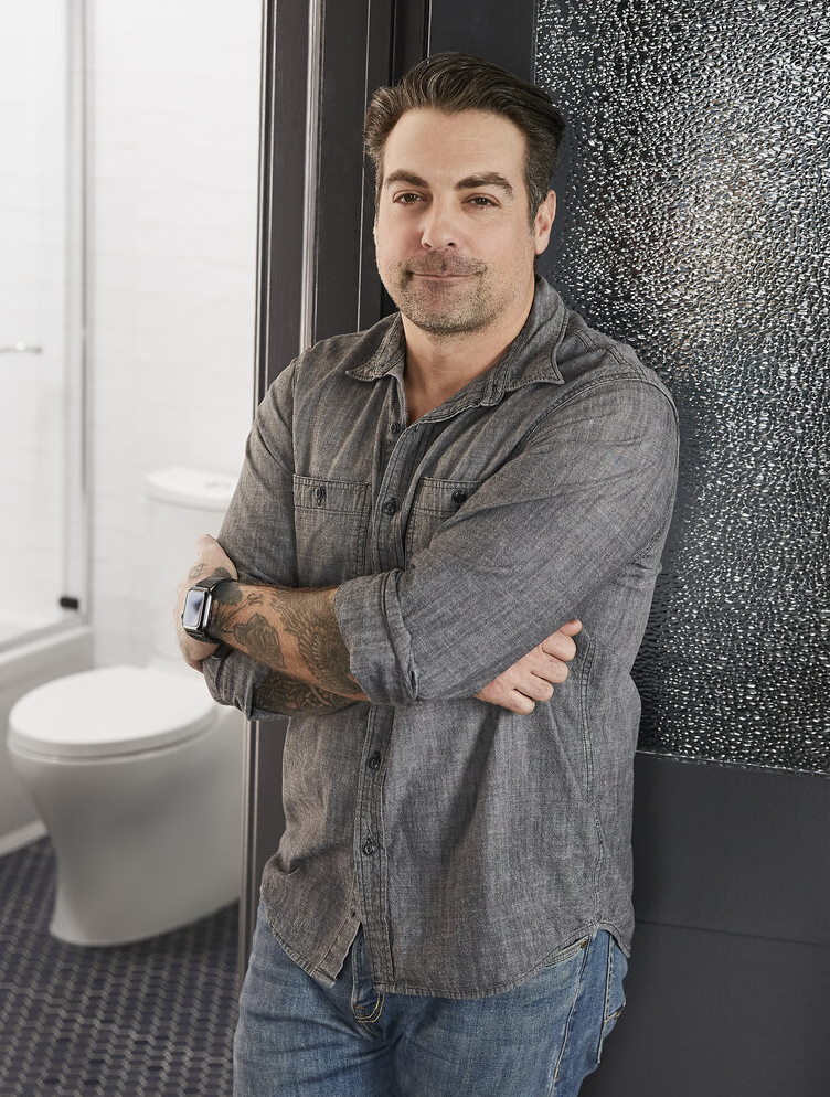 Man in gray shirt leaning against bathroom entry.