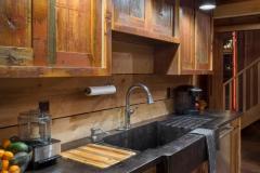 A view of the rustic kitchen in the party barn with wood cabinetry, a black countertop and a backsplash made from horizontal planks of untreated wood.