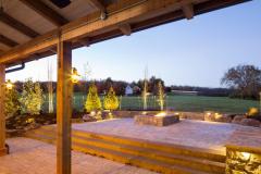 The exterior of the red party barn's covered patio in the evening. A stone accent wall surrounds a fire pit and provides plenty of seating.